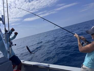 Two anglers actively fishing on a boat, with one reeling in a fish that is jumping out of the water, showcasing an exciting moment on the open sea.