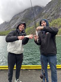 Two anglers proudly displaying their catches, a couple of fish, while fishing in a scenic location surrounded by mountains.