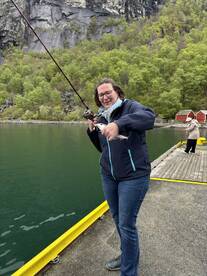 A woman joyfully holding a freshly caught fish while fishing from a dock in a scenic, green landscape.