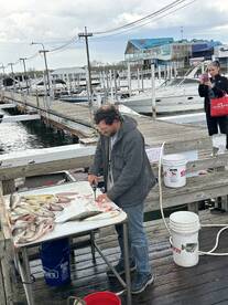 A fisherman filleting a variety of fish on a dock, showcasing a catch that includes several species, with boats visible in the background.