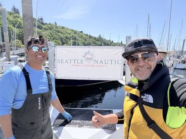 Two smiling individuals pose in front of the Seattle Nautic Fishing & Boating Charter sign, showcasing a sunny day at the marina.