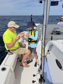 A young angler in a life jacket is learning to handle a fish with the guidance of an adult on a fishing boat, showcasing a moment of mentorship and excitement on the water.