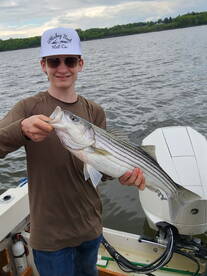 A young angler proudly displaying a large Striped Bass while fishing on a boat, showcasing a successful day on the water.