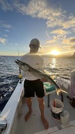 A young angler proudly displaying a Barracuda against a stunning sunset backdrop while fishing on a boat.