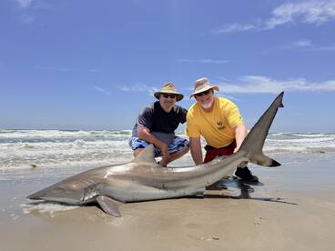 Two anglers proudly displaying a large shark on the beach, showcasing their successful catch during a coastal fishing adventure.
