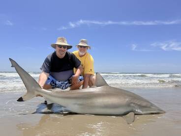 Two anglers proudly displaying a large shark on the beach, showcasing a successful day of fishing in a coastal environment.