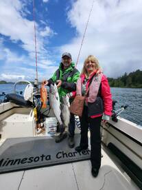 Two anglers proudly displaying their catch, including a large salmon and a rockfish, aboard a charter boat in a scenic fishing environment.