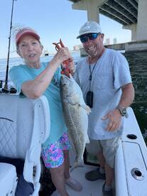 A woman proudly holds a large Black Drum while fishing with a guide under a bridge, showcasing a successful day on the water.