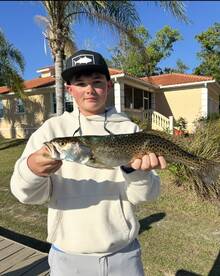 Young angler proudly holding a large Speckled Trout in front of a residential backdrop after a successful fishing trip.