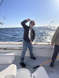 A young angler proudly displaying a striped bass while fishing on a boat, with a clear blue sky and ocean in the background.
