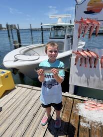 A young angler proudly displaying a fish at the dock, with a boat and a catch board featuring multiple fish in the background.