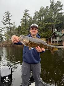 Angler proudly displaying a large Smallmouth Bass while fishing from a boat in a scenic lake surrounded by trees.