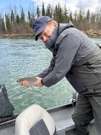 An angler proudly displaying a Rainbow Trout while fishing from a boat in a scenic river setting.