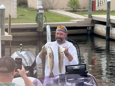 Angler proudly displaying two Redfish while another person captures the moment on a smartphone, showcasing a successful day on the water.