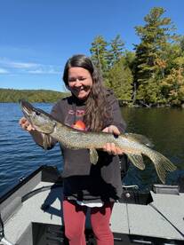 A happy angler proudly displaying a Northern Pike while fishing on a serene lake surrounded by lush greenery.