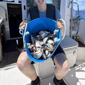 A young angler is smiling while holding a blue bucket filled with various fish aboard a fishing boat, showcasing a successful catch.