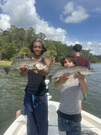 Two young anglers proudly displaying their catches of Speckled Trout while fishing on a boat in a scenic waterway.