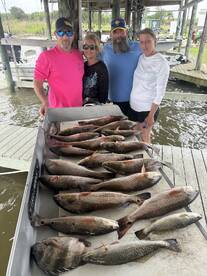 A group of four anglers proudly displaying their catch of various fish, including speckled trout, on a boat's catch board at the dock.