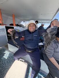 A happy angler proudly displaying two Black Sea Bass on a fishing charter, surrounded by fellow anglers on a boat.