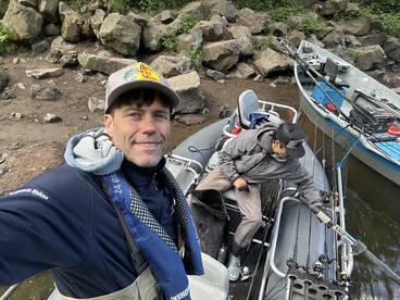 Two anglers preparing their gear on a fishing boat, surrounded by rocky terrain and a calm waterway.