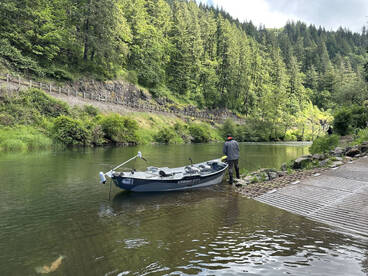 A fisherman stands by a small fishing boat on a serene river, surrounded by lush greenery, preparing to cast his line.