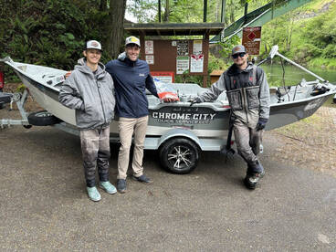Three anglers posing by their fishing boat, 'Chrome City Guide Service,' at a scenic river location, showcasing a friendly atmosphere.