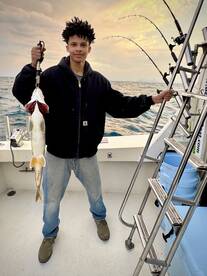 A young angler proudly displaying a large fish on a fishing boat, with rods in the background and a scenic ocean view.