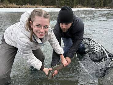 A young angler and an adult are joyfully holding a Rainbow Trout in a river setting, showcasing a successful catch during a fishing trip.
