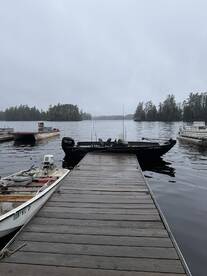 A sleek black fishing boat docked at a serene lake, surrounded by lush trees under a cloudy sky.