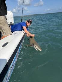 Angler reeling in a shark while fishing from a boat in clear blue waters, showcasing an exciting catch in progress.