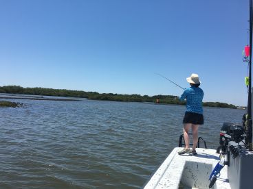 An angler casting a line from a small boat in a serene coastal environment, surrounded by lush greenery.
