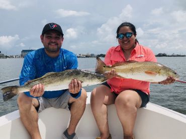 Two anglers proudly displaying their catches, a Speckled Trout and a Redfish, while enjoying a sunny day on the water.