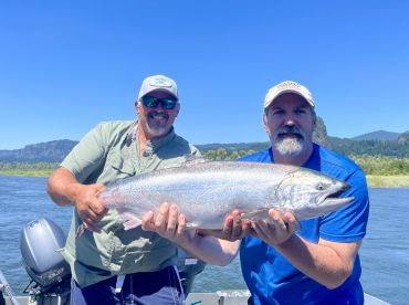 Two anglers proudly displaying a large Chinook Salmon while fishing on a river, surrounded by scenic mountains.