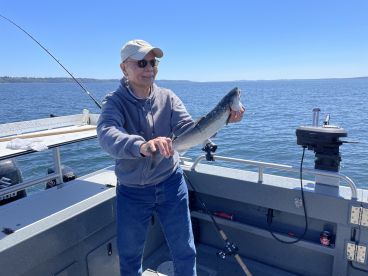 An older angler proudly displaying a freshly caught fish while fishing on a boat, surrounded by a scenic water backdrop.