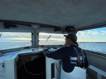 A captain at the helm of a fishing boat, navigating through open waters during a scenic sunrise.