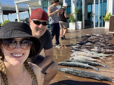 A group of anglers poses near a display of various fish, including Mahi Mahi and Snapper, on a sunny day at the dock.