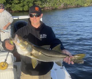 Angler proudly displaying a large Snook while fishing in a scenic waterway, with another fisherman visible in the background.