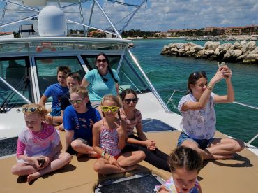 A group of children and adults enjoying a sunny day on a boat, capturing memories with smiles and playful poses against a vibrant coastal backdrop.
