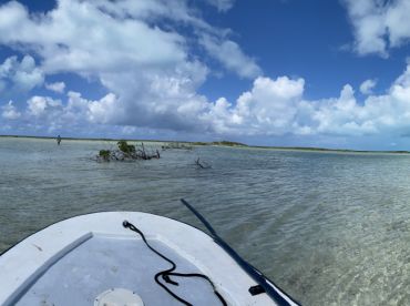 A scenic view from the bow of a boat in shallow waters, showcasing a fishing environment with a person wading in the distance.