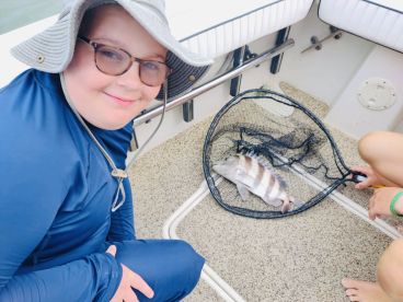 A young angler smiles while sitting on a boat next to a striped fish in a net, showcasing a successful fishing trip.