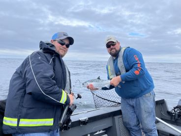 Two anglers proudly displaying their catch, a Silver Salmon, while fishing on a boat in open waters.