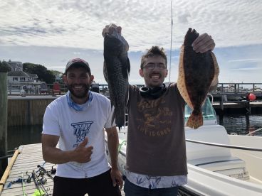 Two anglers proudly displaying their catch of a Black Sea Bass and a Flounder at the dock after a successful fishing trip.