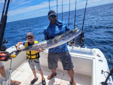 A young angler and an adult proudly displaying a large Kingfish aboard a fishing boat in open waters.