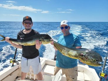 Two anglers proudly displaying their large Mahi Mahi catches while aboard a fishing boat in clear blue waters.