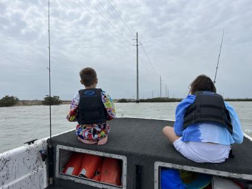Two young anglers sitting on the back of a boat, enjoying a fishing trip in a calm waterway, surrounded by mangroves and power lines.