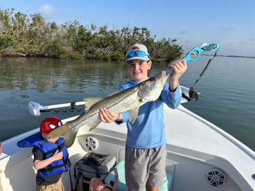A young angler proudly displaying a Snook caught while fishing on a boat, with a scenic backdrop of mangroves.