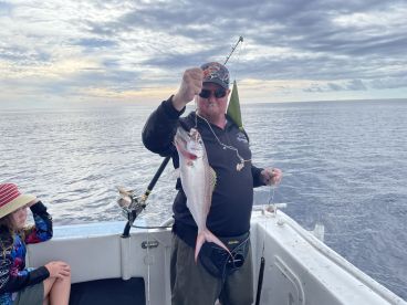 An angler proudly displaying a Snapper caught during a deep-sea fishing trip, with a young child observing on the boat.