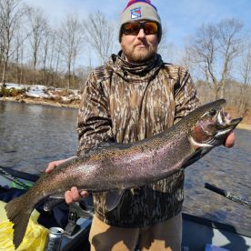Angler displaying a large Rainbow Trout while fishing in a scenic river setting during early spring.