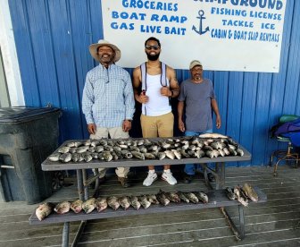 Three anglers proudly displaying their catch of various fish on a table at a fishing dock, showcasing a successful day on the water.