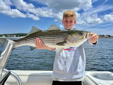 A young angler proudly displaying a large Striped Bass while fishing on a boat, showcasing a beautiful day on the water.